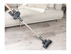 A person vacuuming a light grey wooden floor with a grey cordless stick vacuum cleaner.