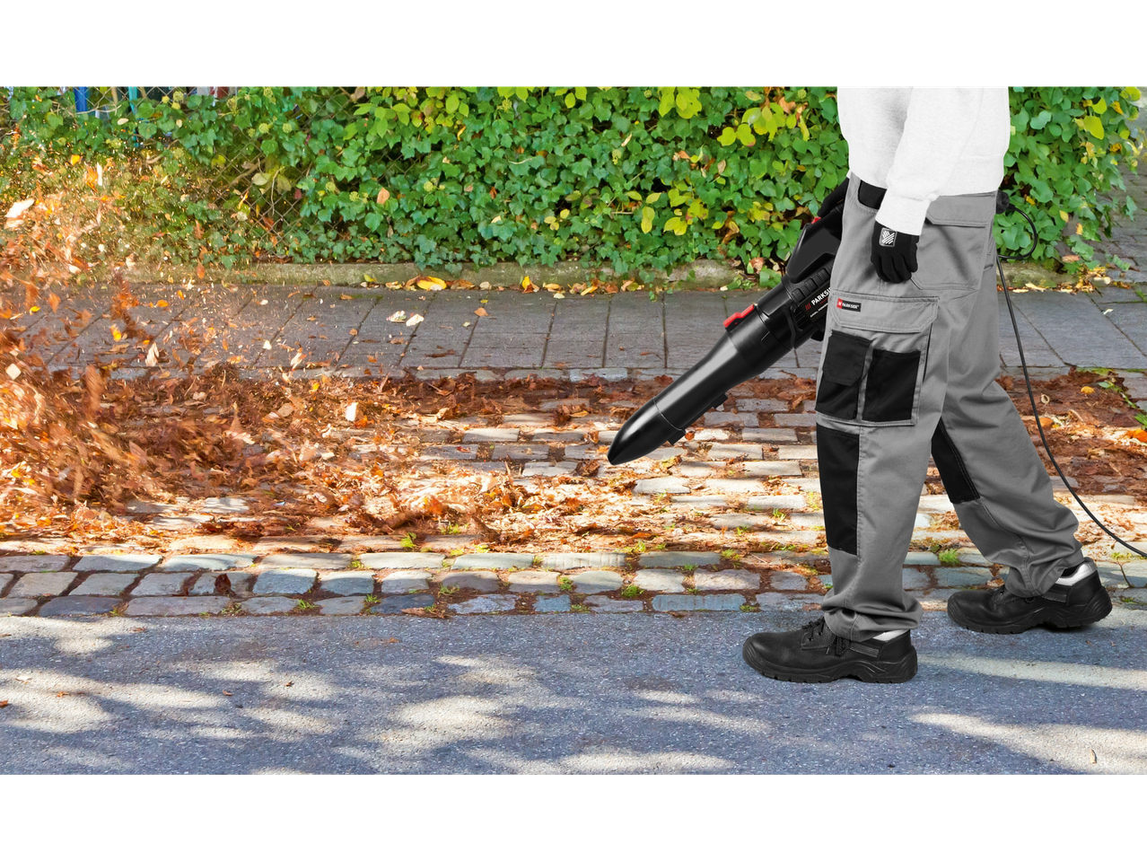 Ένα άτομο χρησιμοποιεί φυσητήρα φύλλων Parkside για να καθαρίσει τα φθινοπωρινά φύλλα από έναν δρόμο με πλακόστρωτο.