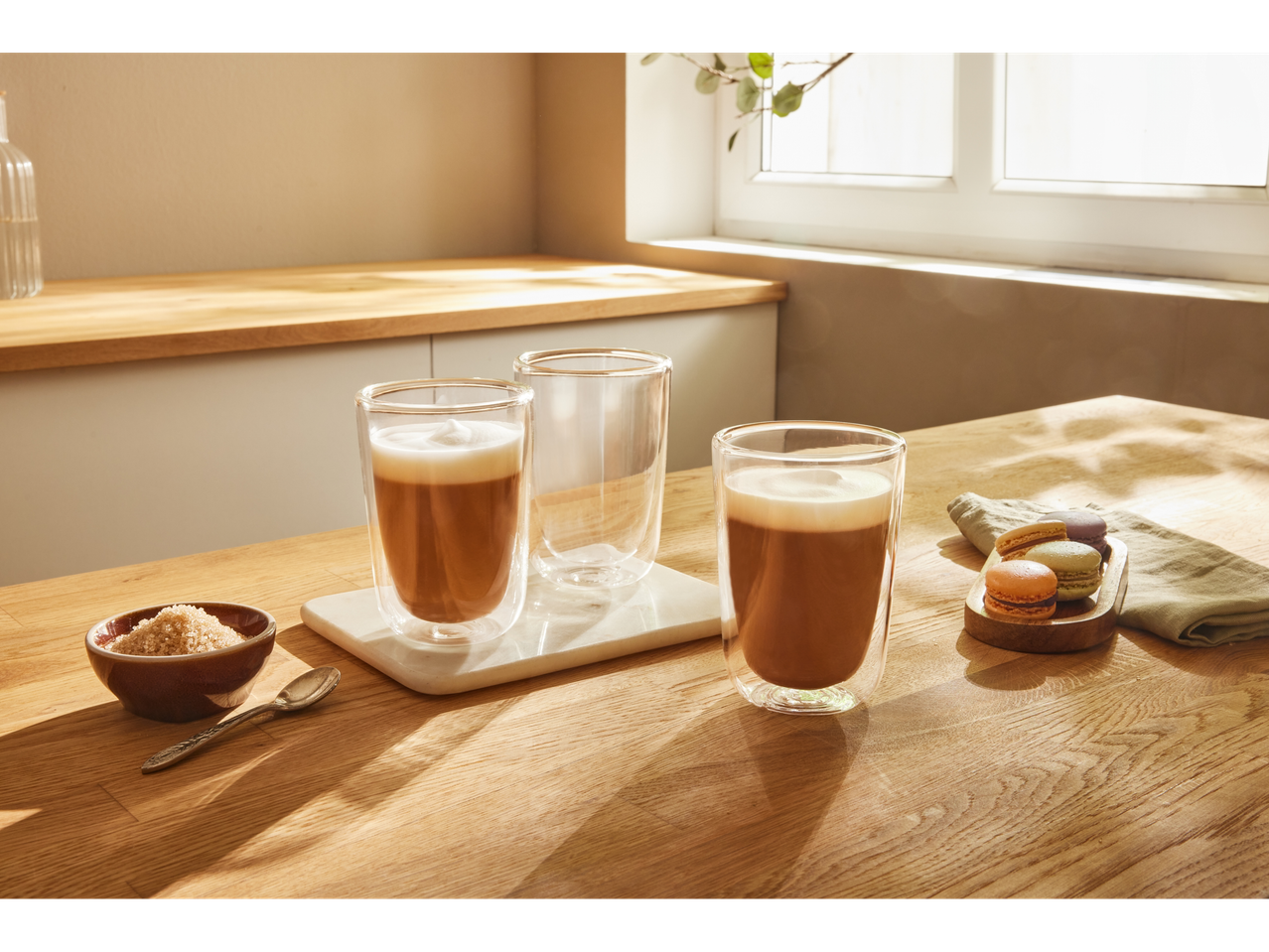 Three latte glasses on a wooden table with macarons and sugar.