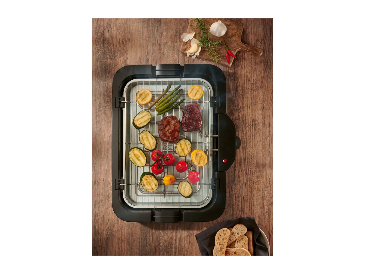 Electric grill with grilled vegetables and meat, garlic, rosemary, and bread on a wooden table.