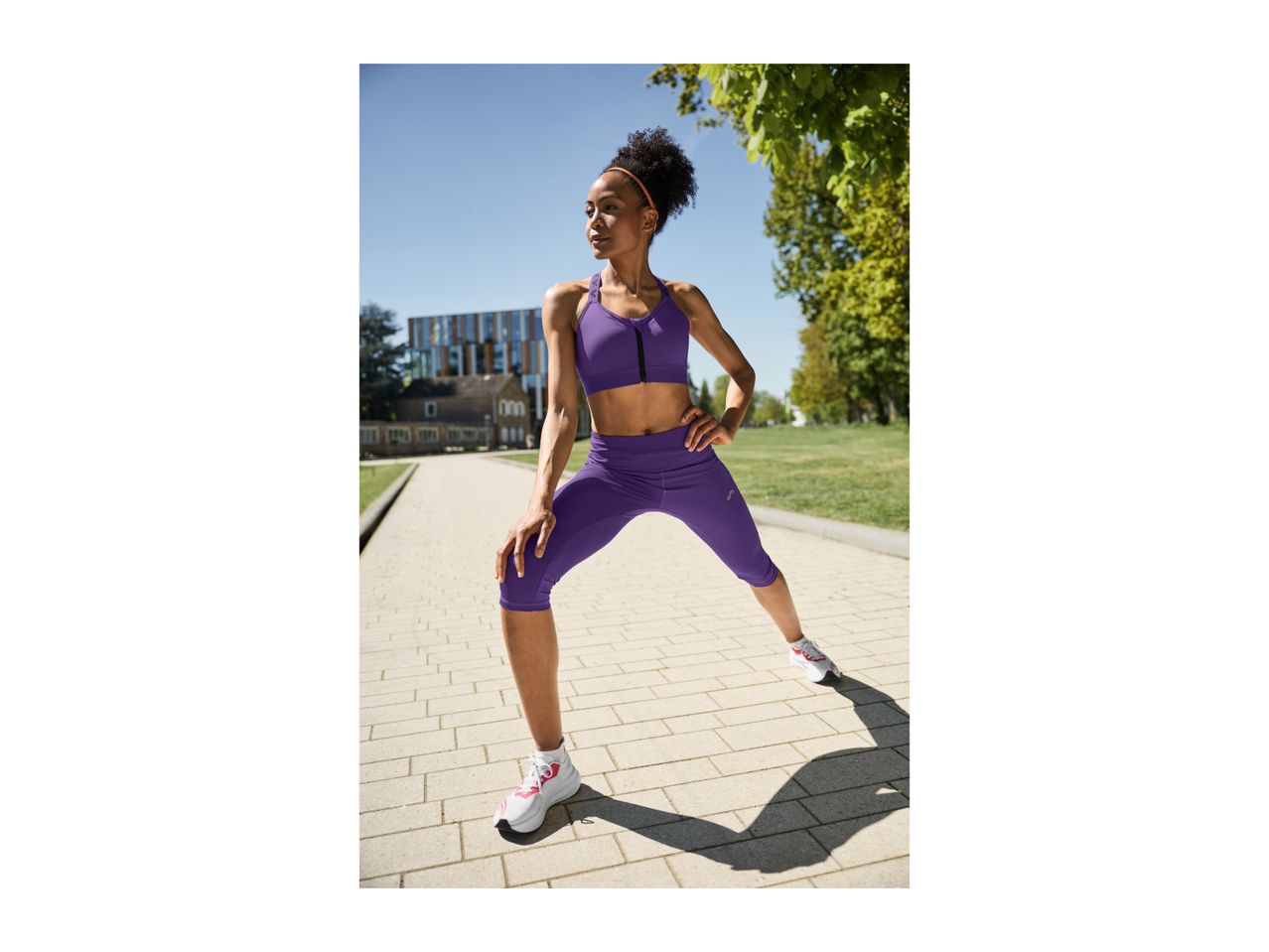 Woman in purple sports bra and capri leggings stretching outdoors on a sunny day.