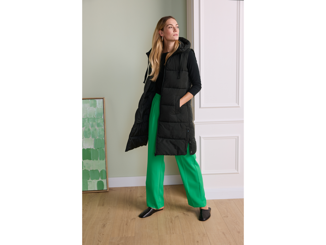 Woman in a long black puffer vest, black long-sleeve top, and bright green trousers.