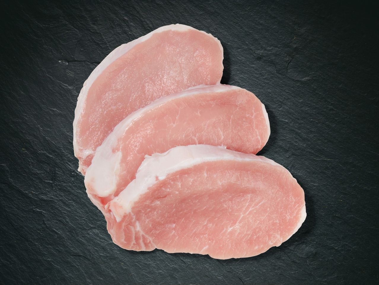 Drei Schweinekoteletts auf dunklem Untergrund.