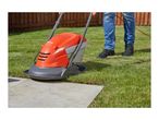 A person in jeans and black shoes uses an orange and grey hover lawnmower on a green lawn next to a patio.