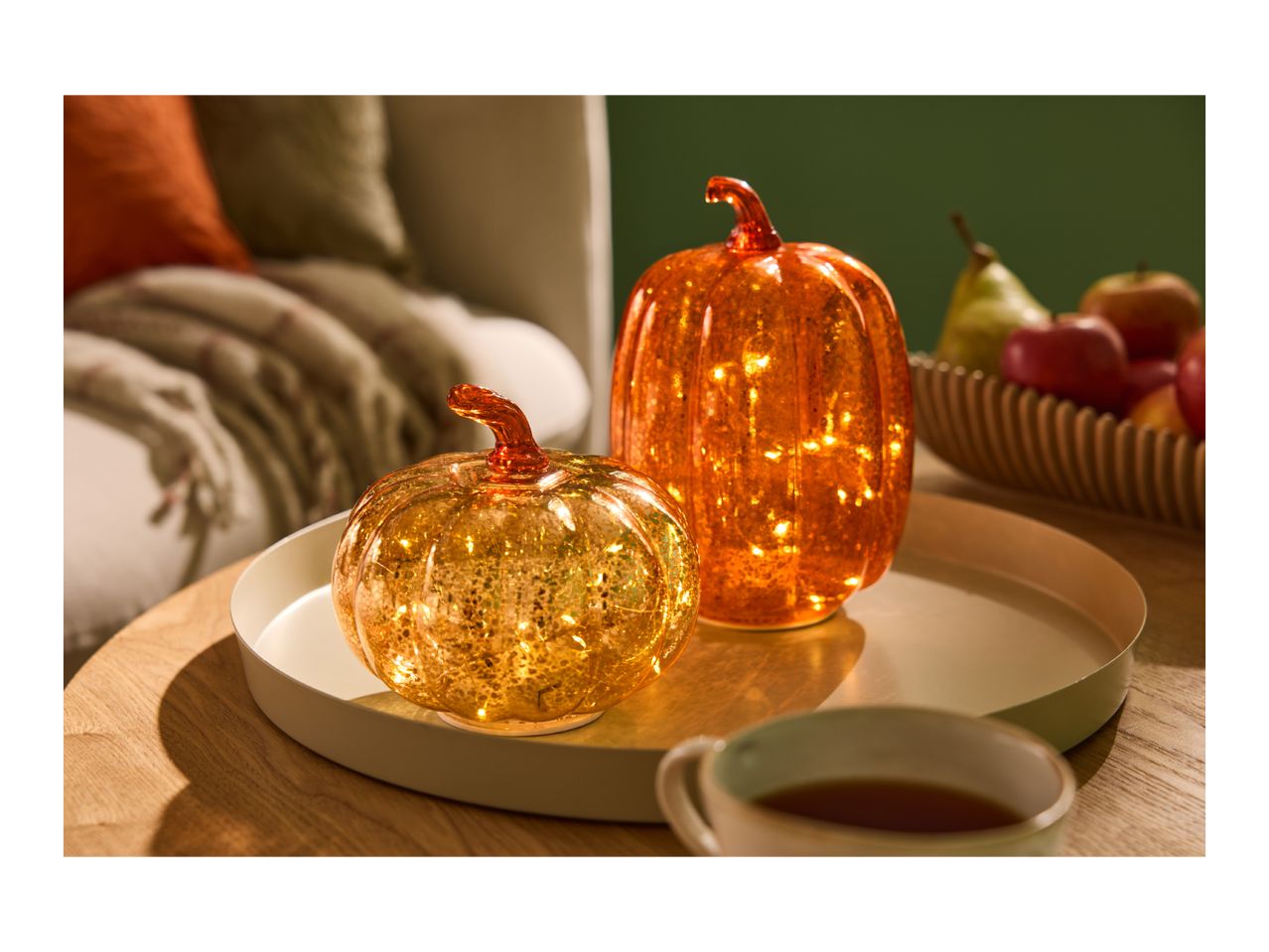 Two illuminated glass pumpkins on a tray, with a cup of tea and fruit in the background.