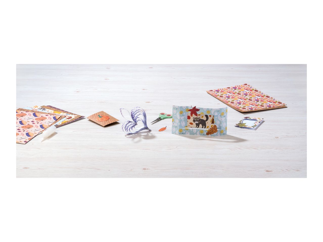 Craft supplies including patterned paper, scissors, glue, and a pop-up card on a wooden table.