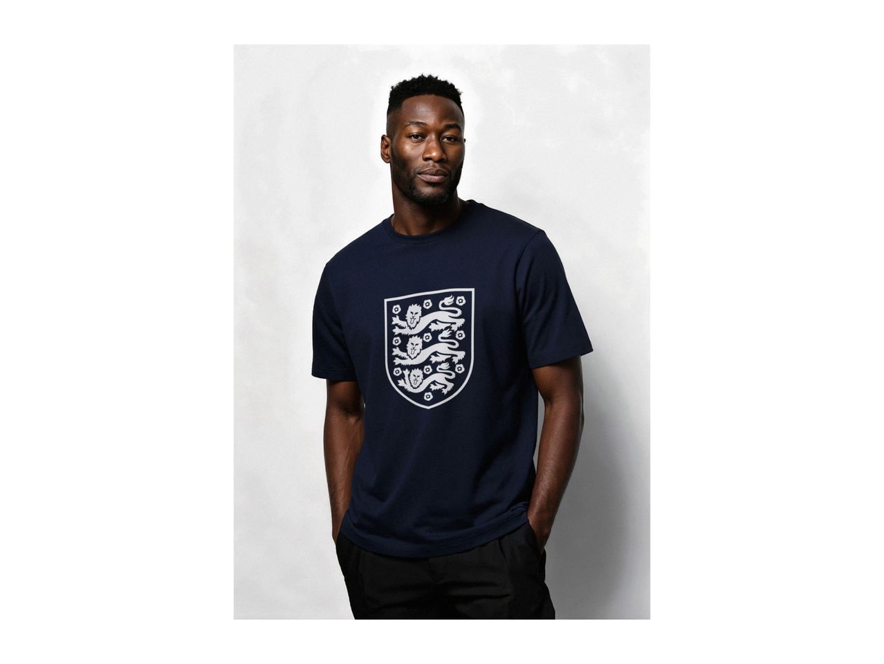 Man wearing a navy blue t-shirt with a white shield emblem, hands in pockets.