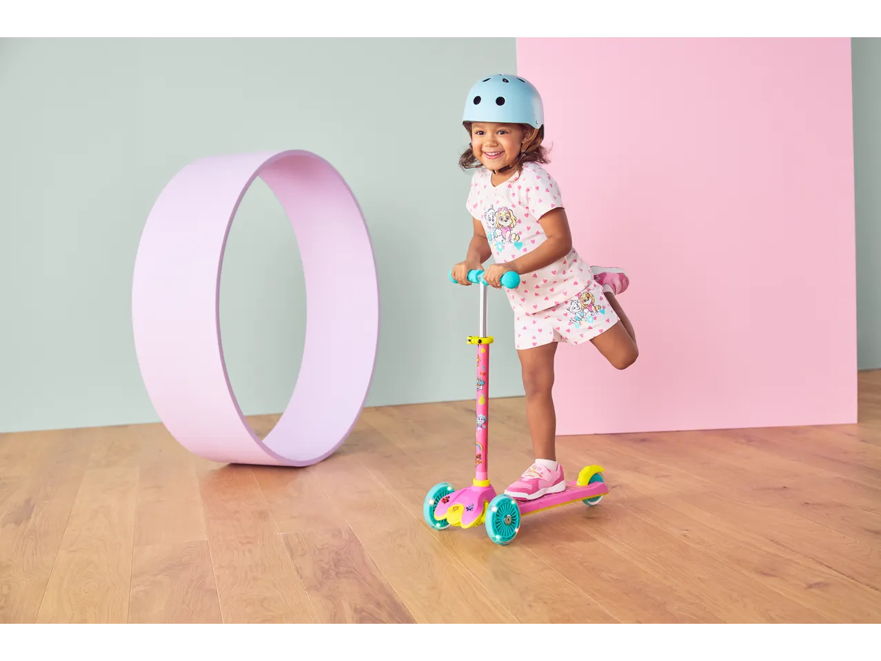 Smiling girl in a blue helmet on a pink Lupilu Tri-Scooter with LED wheels.