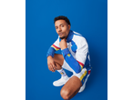 Man in a white, blue, red, and yellow tracksuit, shorts, and socks with a logo, crouching.