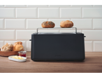 Black toaster with two bread rolls on top, with more rolls, butter, and jam nearby.