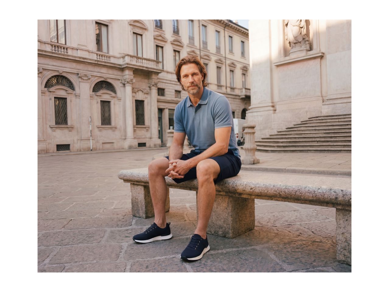 Man in a blue polo shirt and shorts sitting on a stone bench in a city square.