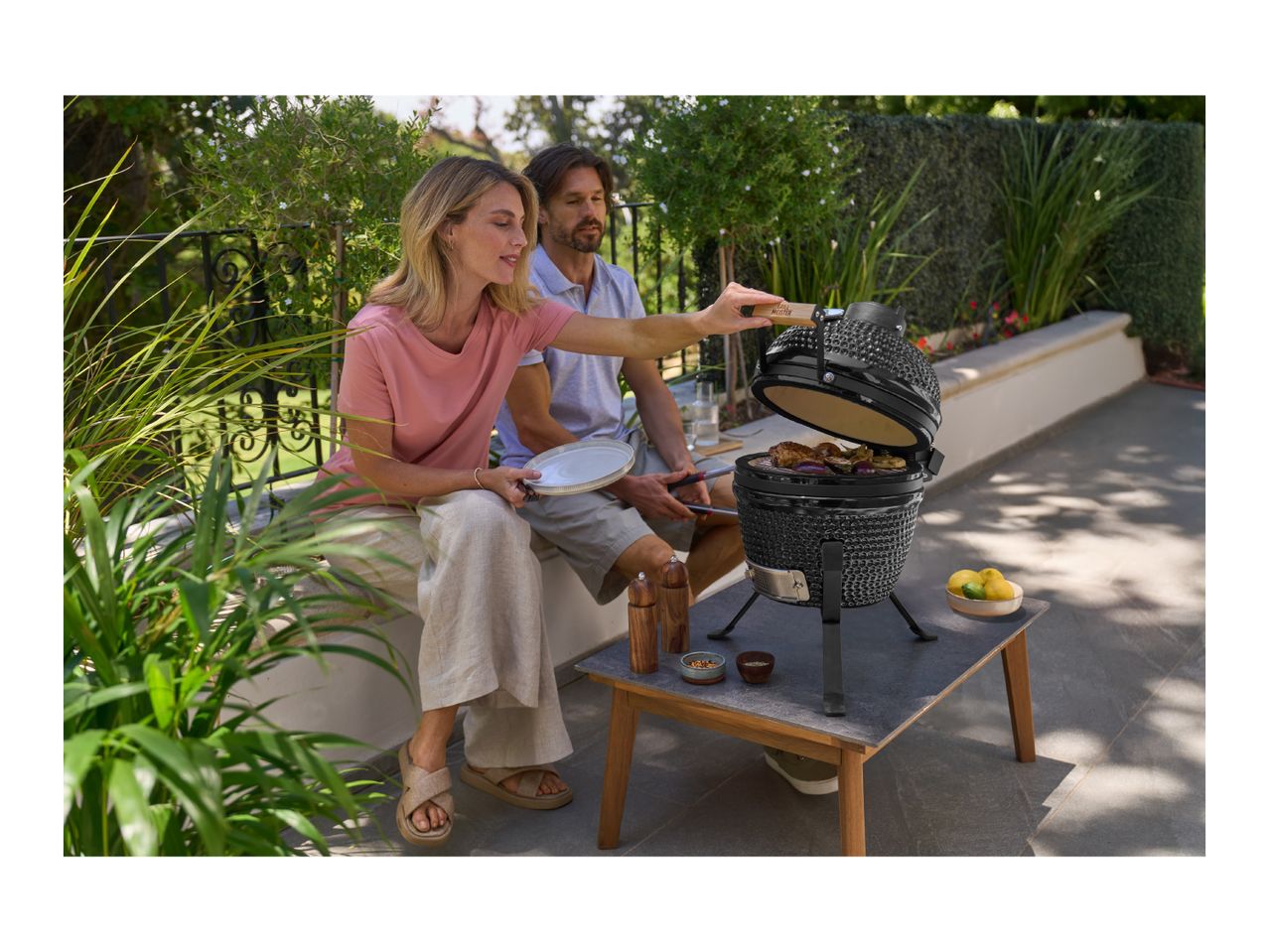 Couple grilling on a small black ceramic barbecue, enjoying an outdoor meal on a sunny patio.