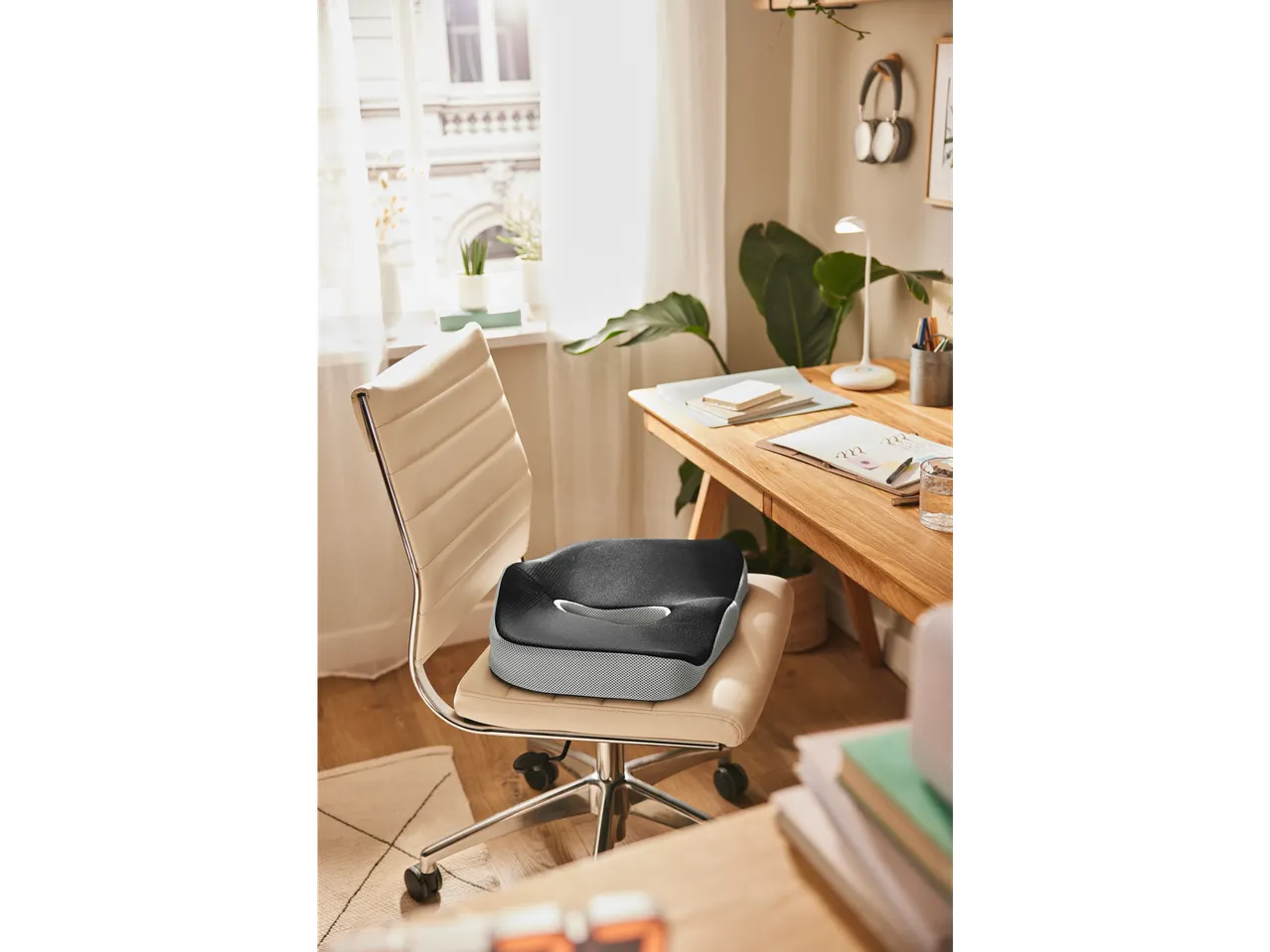 A black and gray memory foam seat cushion sits on a beige office chair in a sunlit home office.