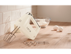 A cream-colored hand mixer with attachments, a bowl of flour, and two eggs on a wooden counter.