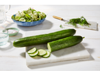 Two fresh cucumbers, one whole and one sliced, on a white marble board with a salad in the background.