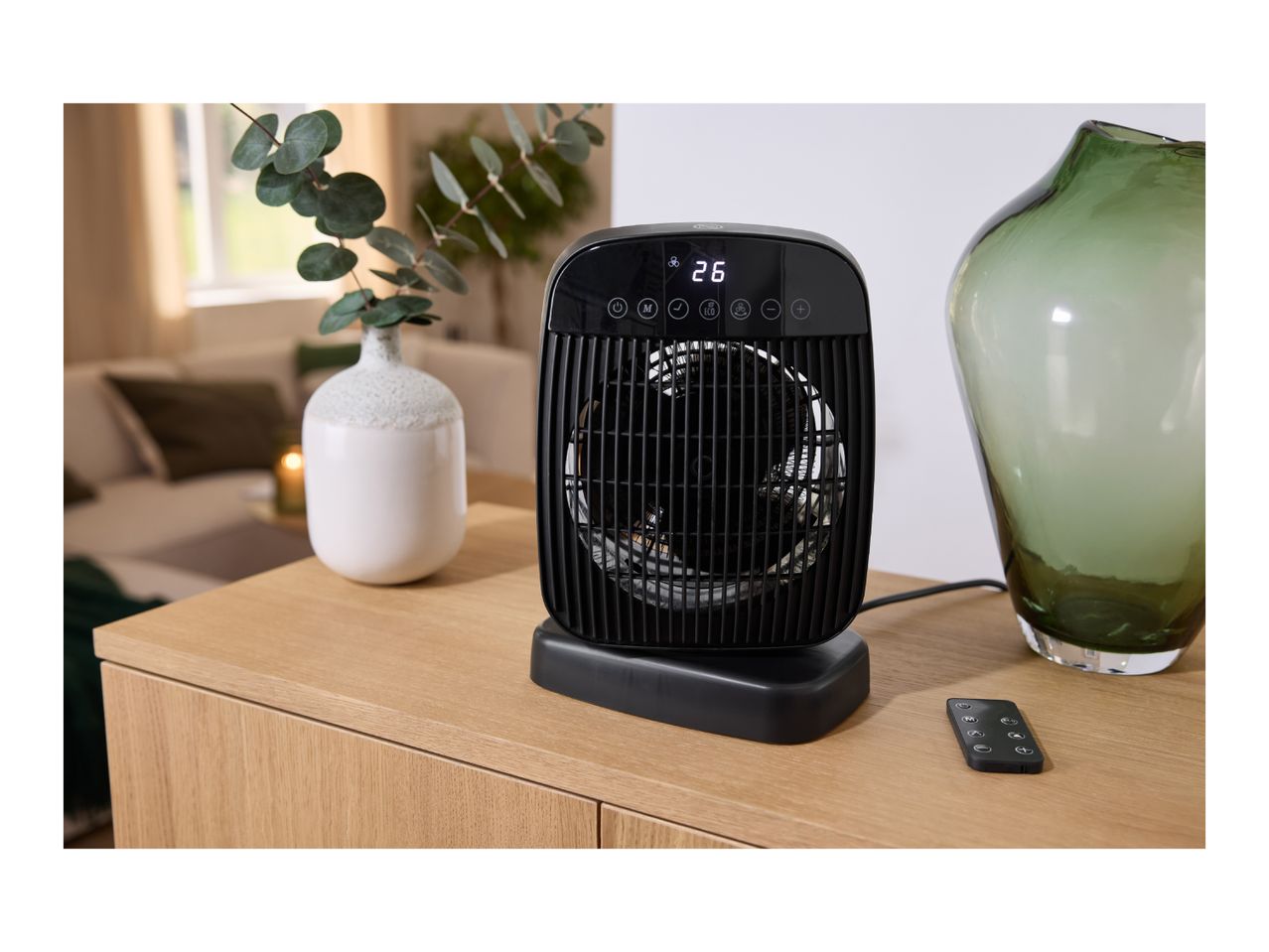 A black ceramic fan heater with a remote control on a wooden sideboard.