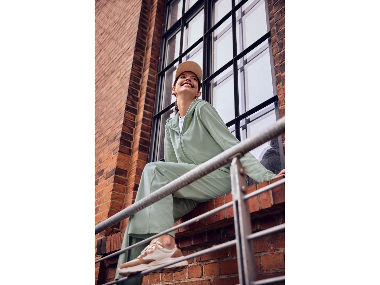 Donna sorridente in tuta verde menta e scarpe da ginnastica beige seduta su un muro di mattoni.