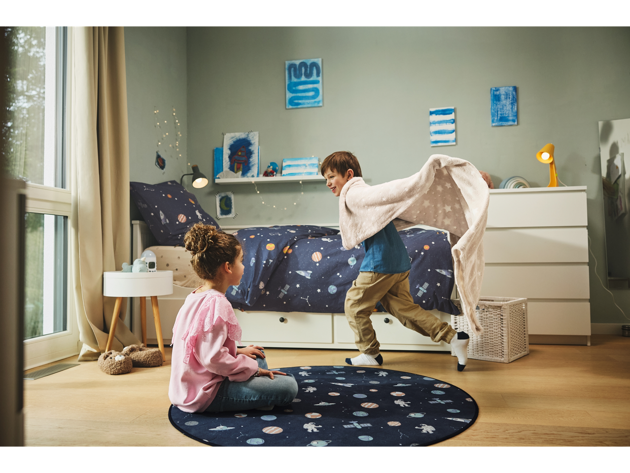 Two children play in a space-themed bedroom with matching rug and blankets.