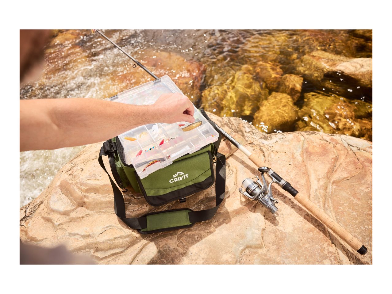 A person selecting a lure from a tackle box, with a fishing rod and reel nearby on rocks by a river.