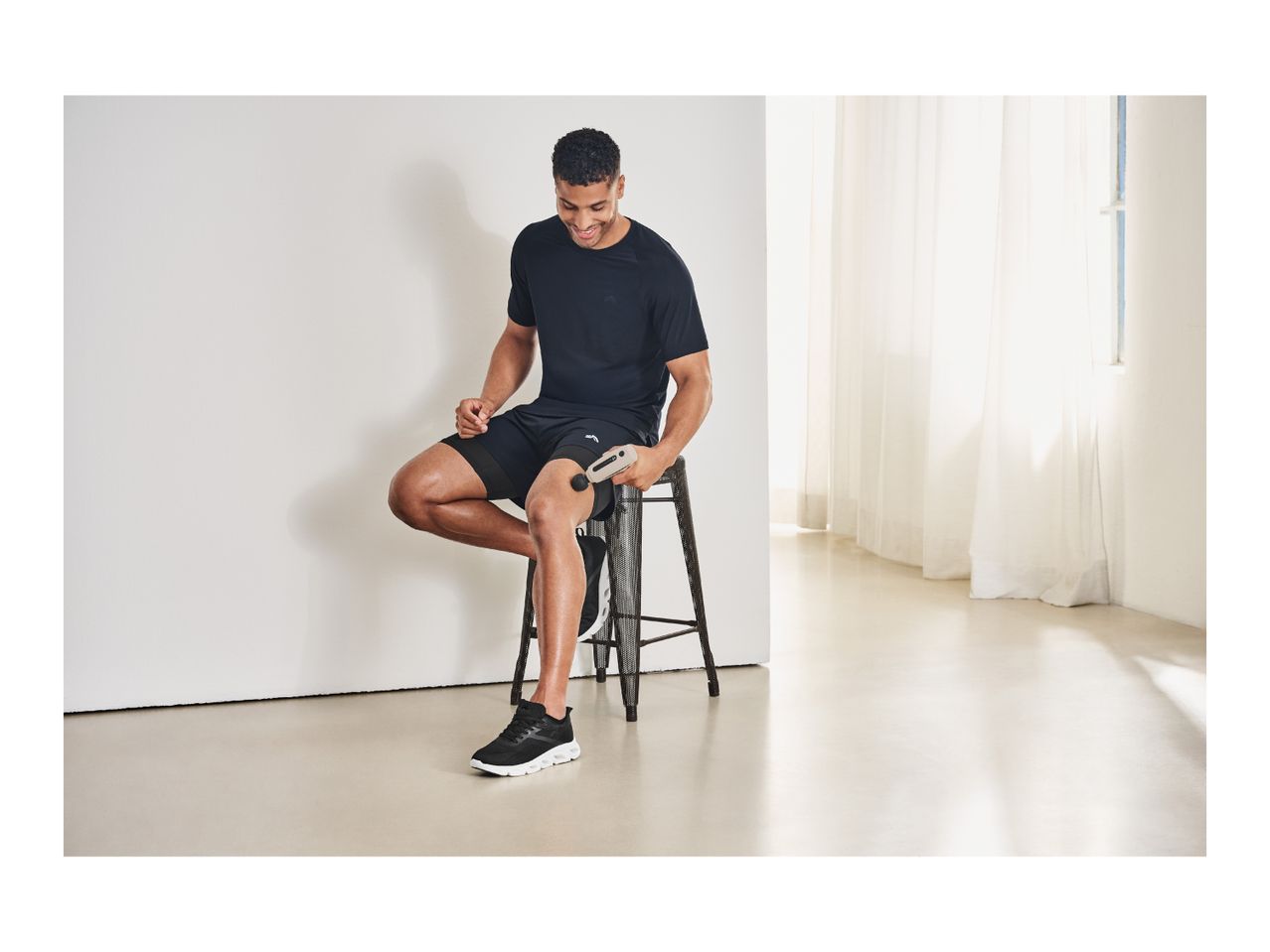 Man in black sportswear using a massage gun on his thigh while sitting on a stool.