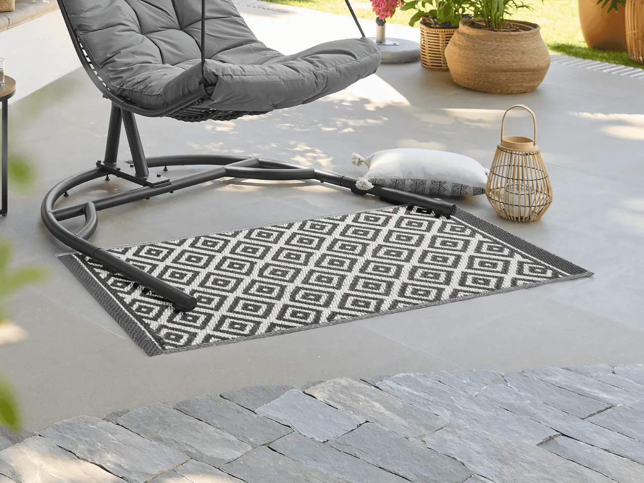 Grauer Hängesessel mit Kissen und schwarz-weißem Outdoor-Teppich auf einer Terrasse.