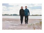Man and woman in outdoor jackets and convertible trousers walking on a beach.