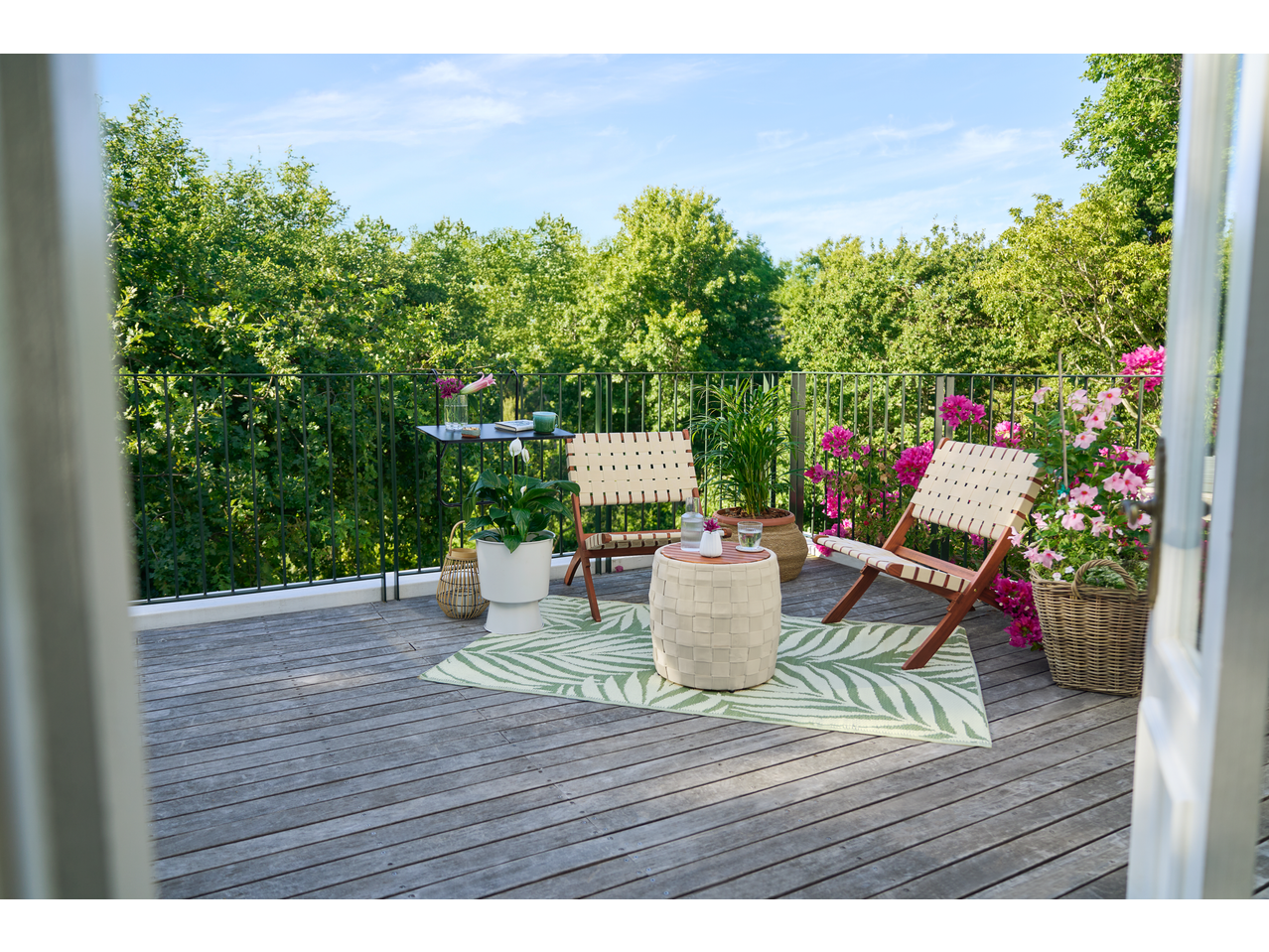 Varanda com cadeiras de madeira e tecido, mesa pequena, tapete verde e vasos de plantas.