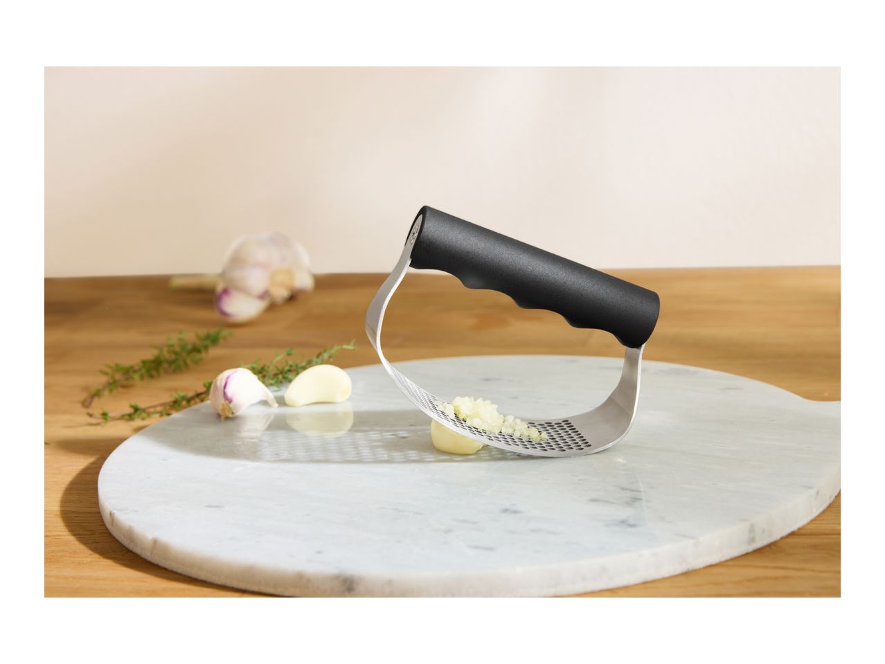 Garlic press with crushed garlic, garlic cloves, and thyme on a marble board.