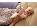 A baby in a floral sleep sack holding a wooden and fabric teether, smiling on a blanket.