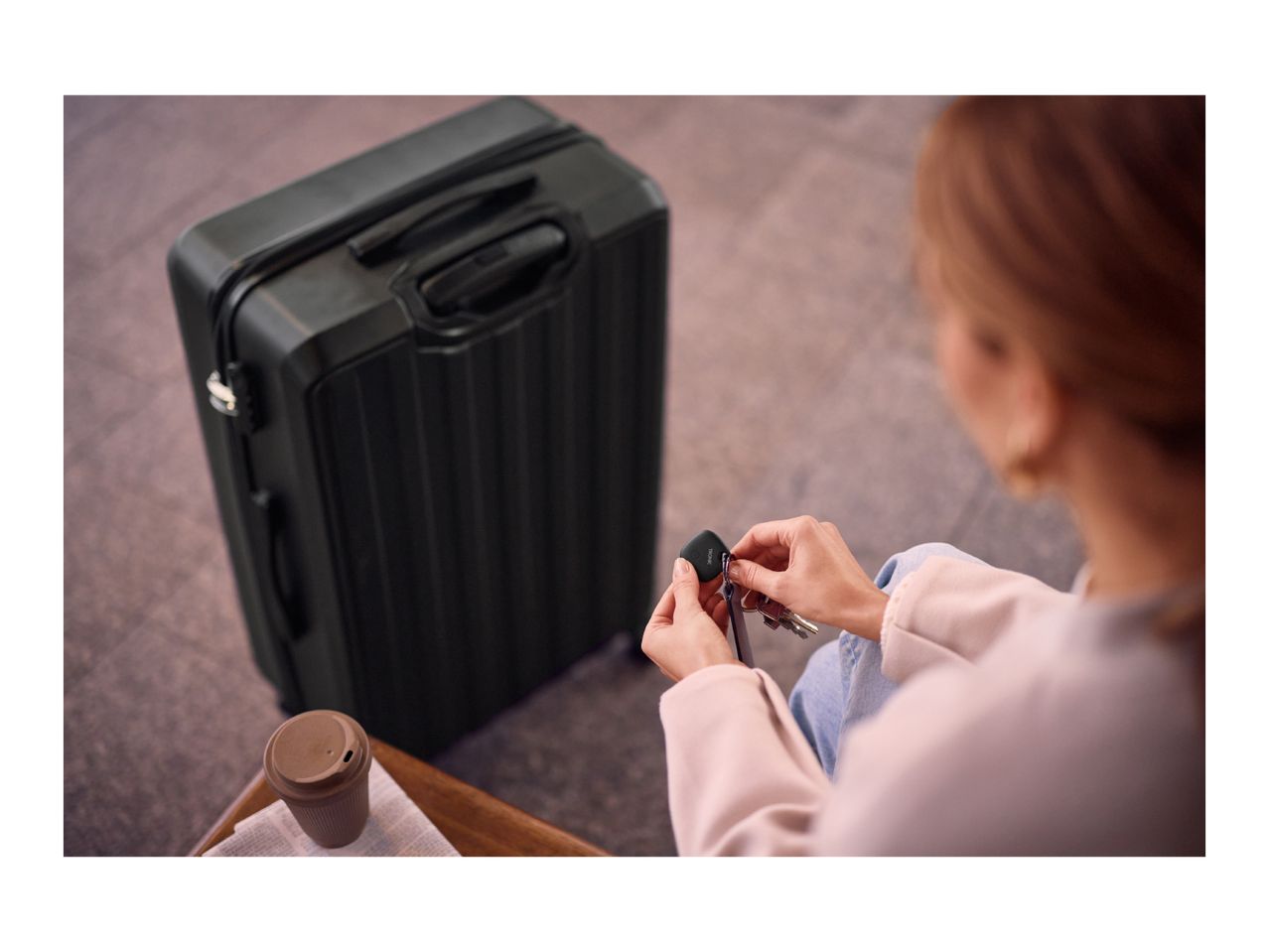 Woman attaching a key finder to keys next to a suitcase and a reusable coffee cup.