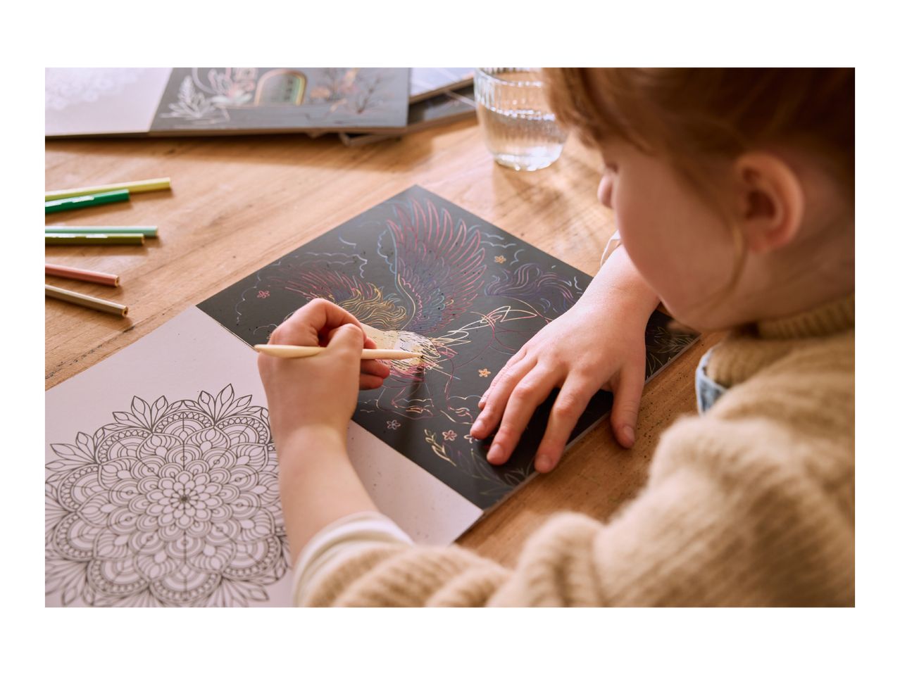 Child scratching a colorful phoenix design on a scratch art book, with a mandala coloring page nearby.