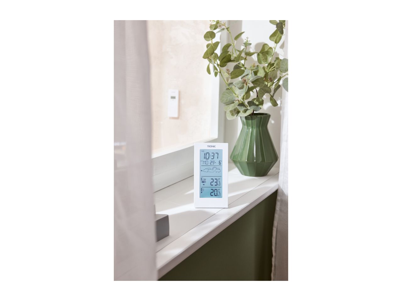 Digital weather station displaying time, date, and temperature, next to a green vase with plants.