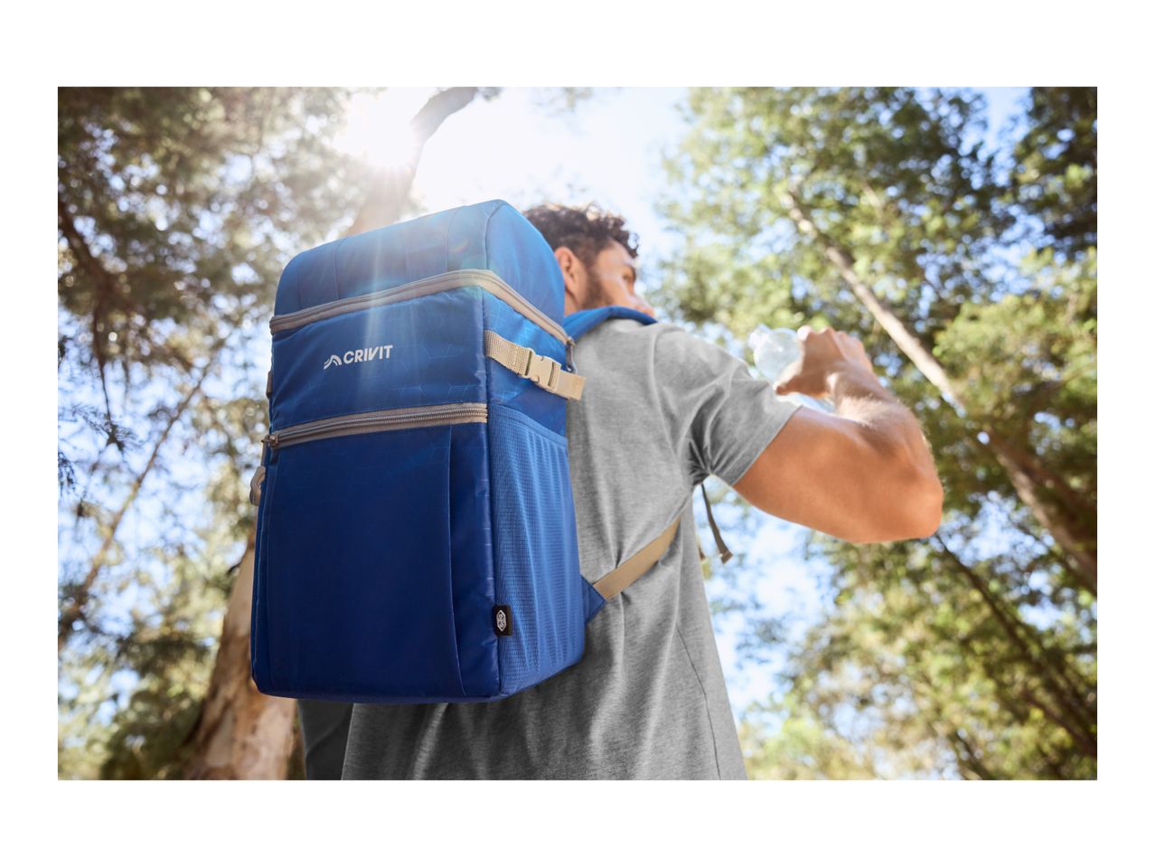 Man wearing a blue cooler backpack, drinking water in a forest setting.