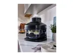 Black food processor with ingredients inside, on a kitchen counter with a plant and plates.