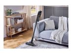 Cordless stick vacuum cleaner on a patterned rug in a living room with a sofa and wooden shelf.
