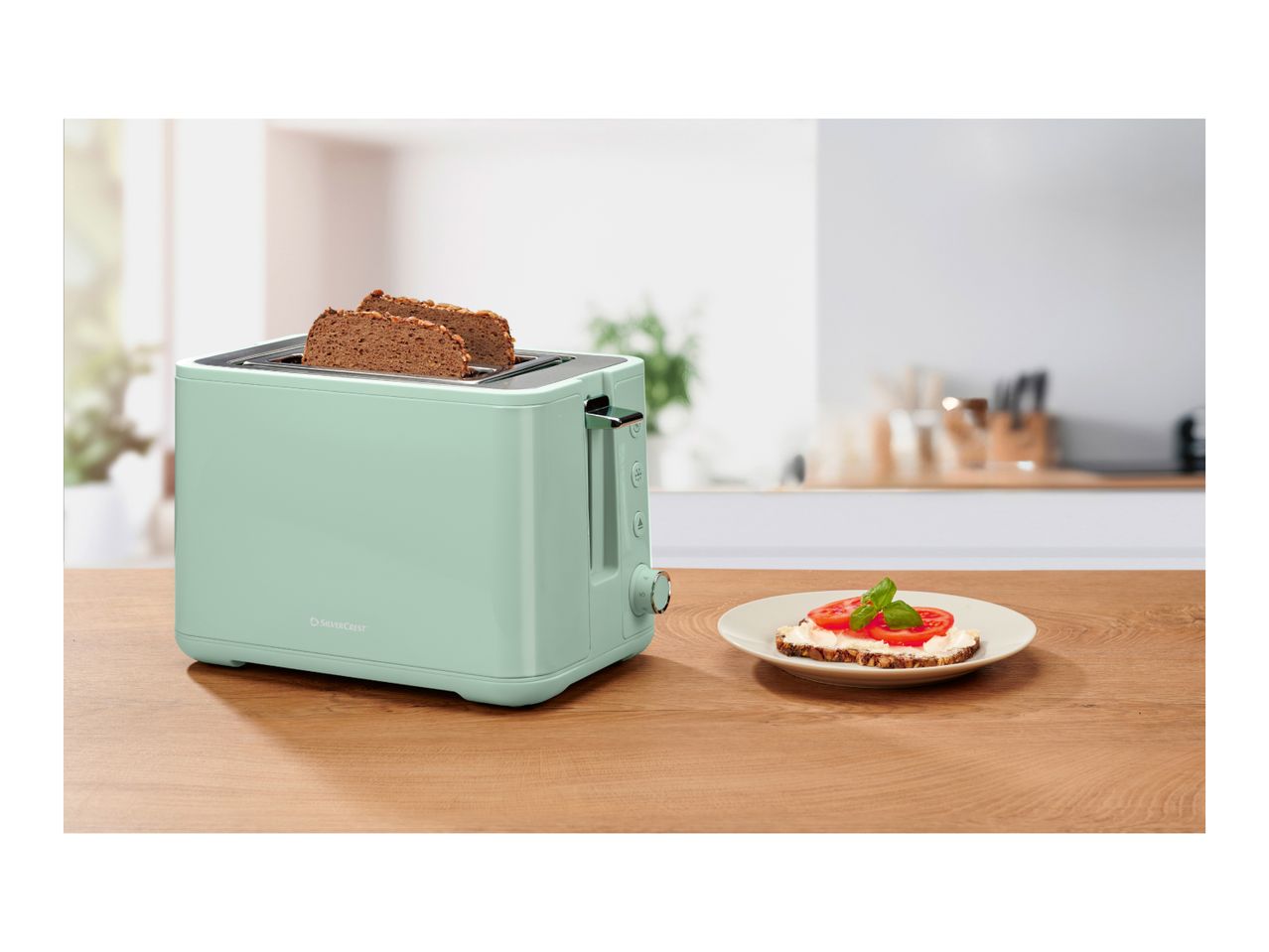 Mint green toaster with two slices of bread, next to a plate with toast and tomato.