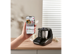 Mão segurando smartphone com app Monsieur Cuisine, ao lado de um SILVERCREST® KITCHEN TOOLS Monsieur Cuisine “Compact”.