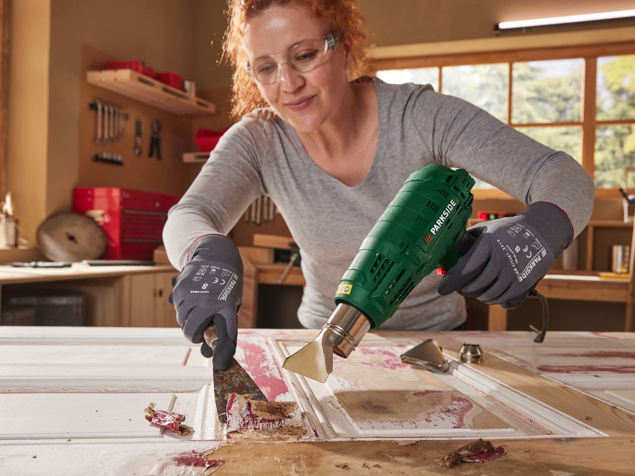 A woman uses a Parkside heat gun and scraper to remove old paint from a door.