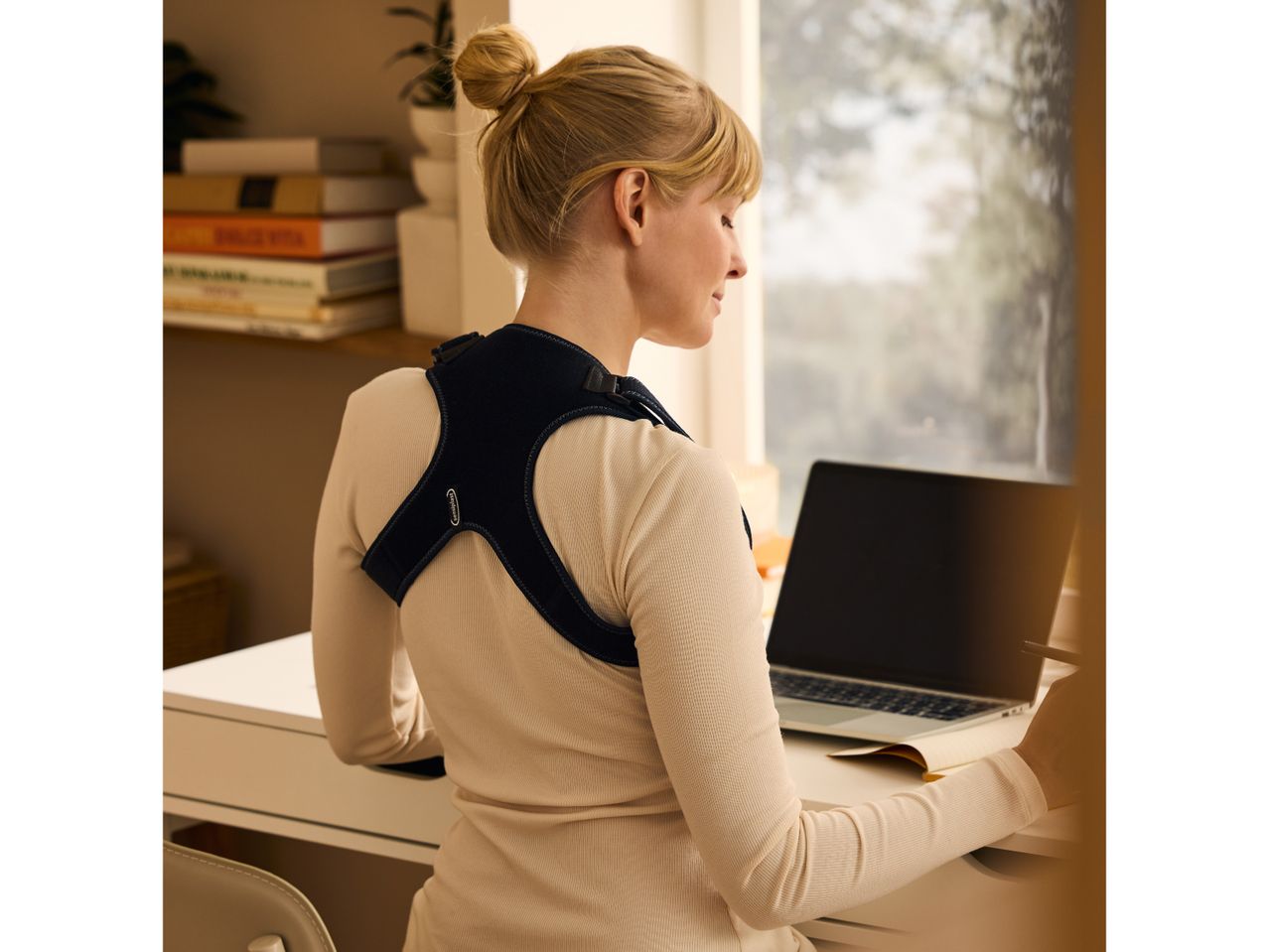 Woman wearing a posture corrector, working on a laptop at a desk.