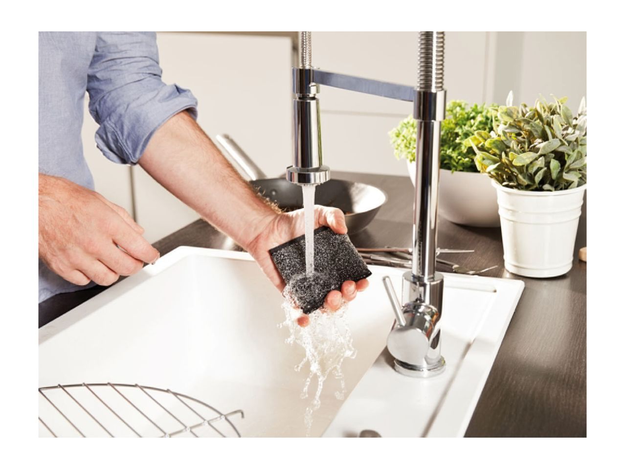 Man rinsing a dark sponge under a modern kitchen faucet in a white sink.