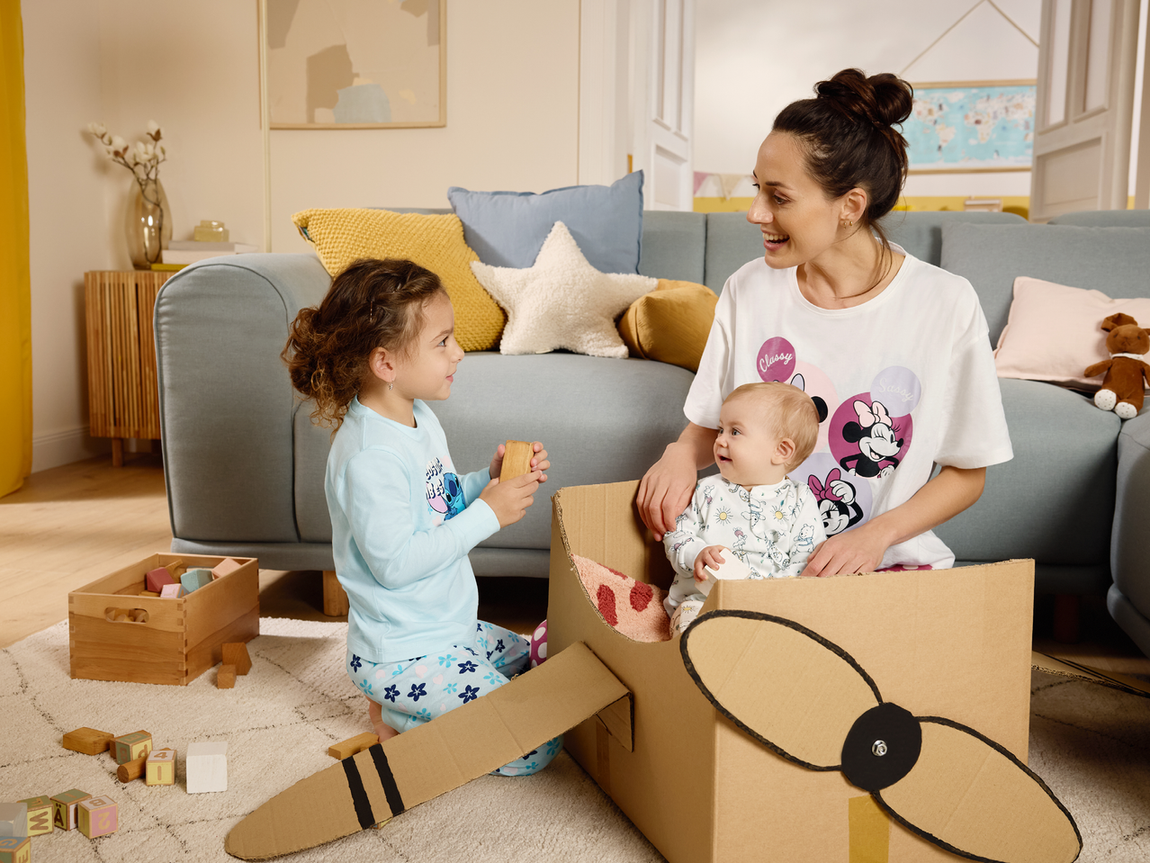 Майка и две деца си играят с картонен самолет и дървени блокчета в хола.