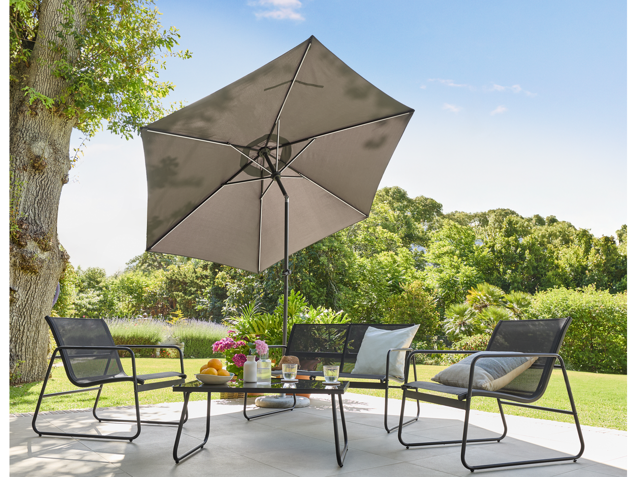 Outdoor patio set with a grey umbrella, black chairs, and a coffee table with drinks and fruit.