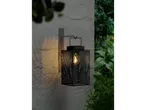 Black outdoor lantern with geometric pattern, mounted on a wall, surrounded by greenery.