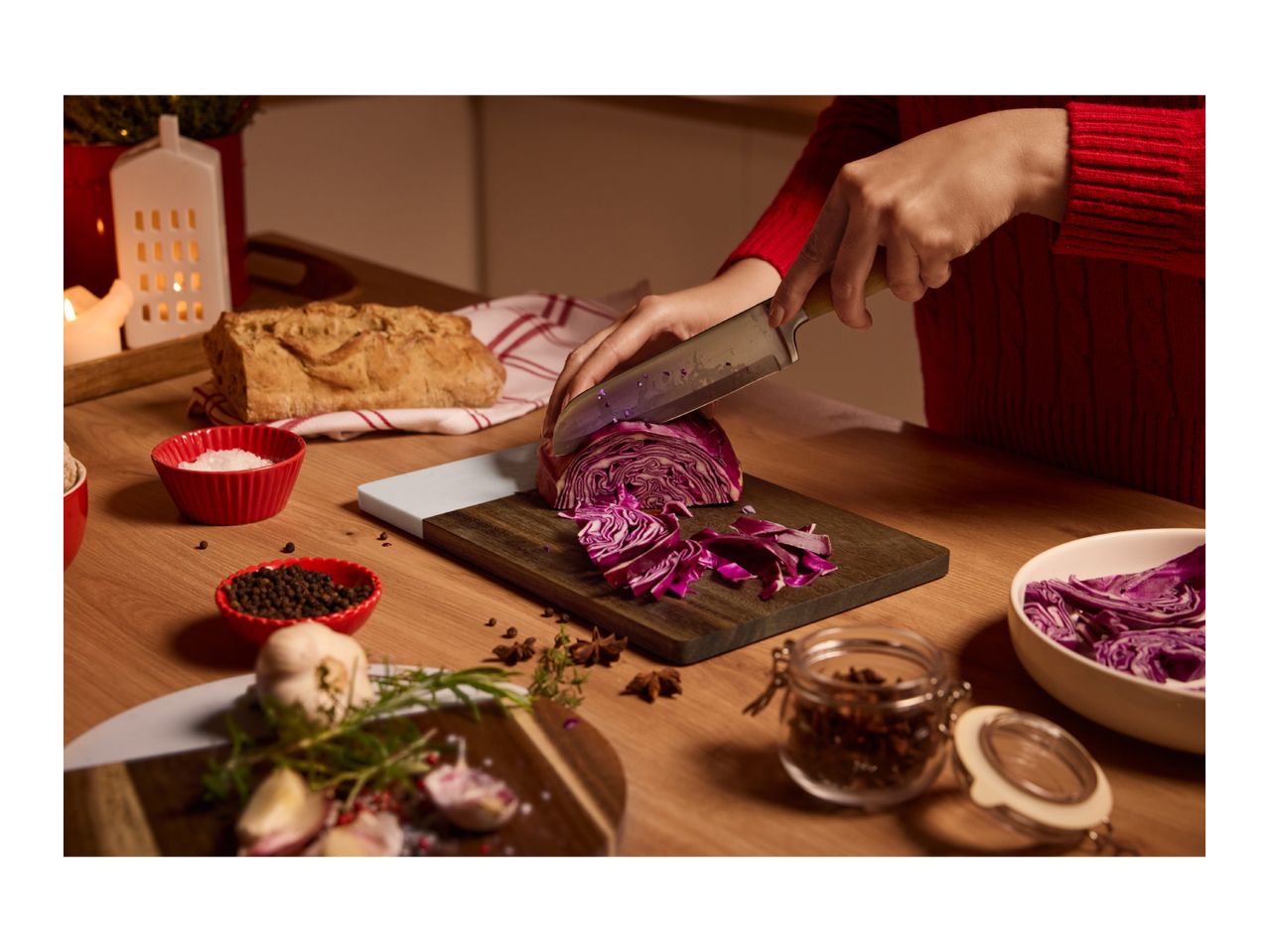 Person chopping red cabbage on a wooden board, surrounded by spices and bread.