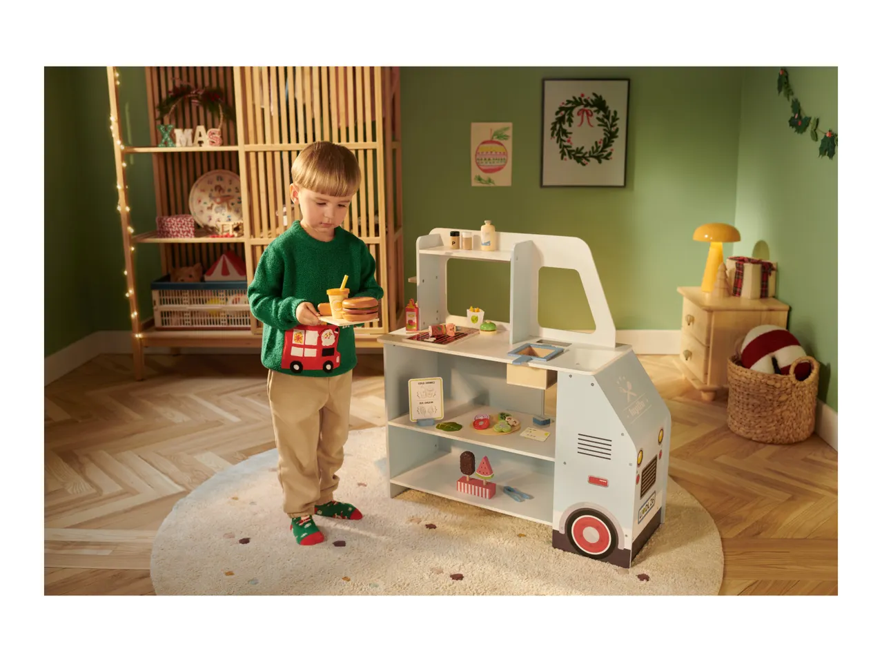 Child playing with a toy food truck, holding a burger and drink, in a festive room.