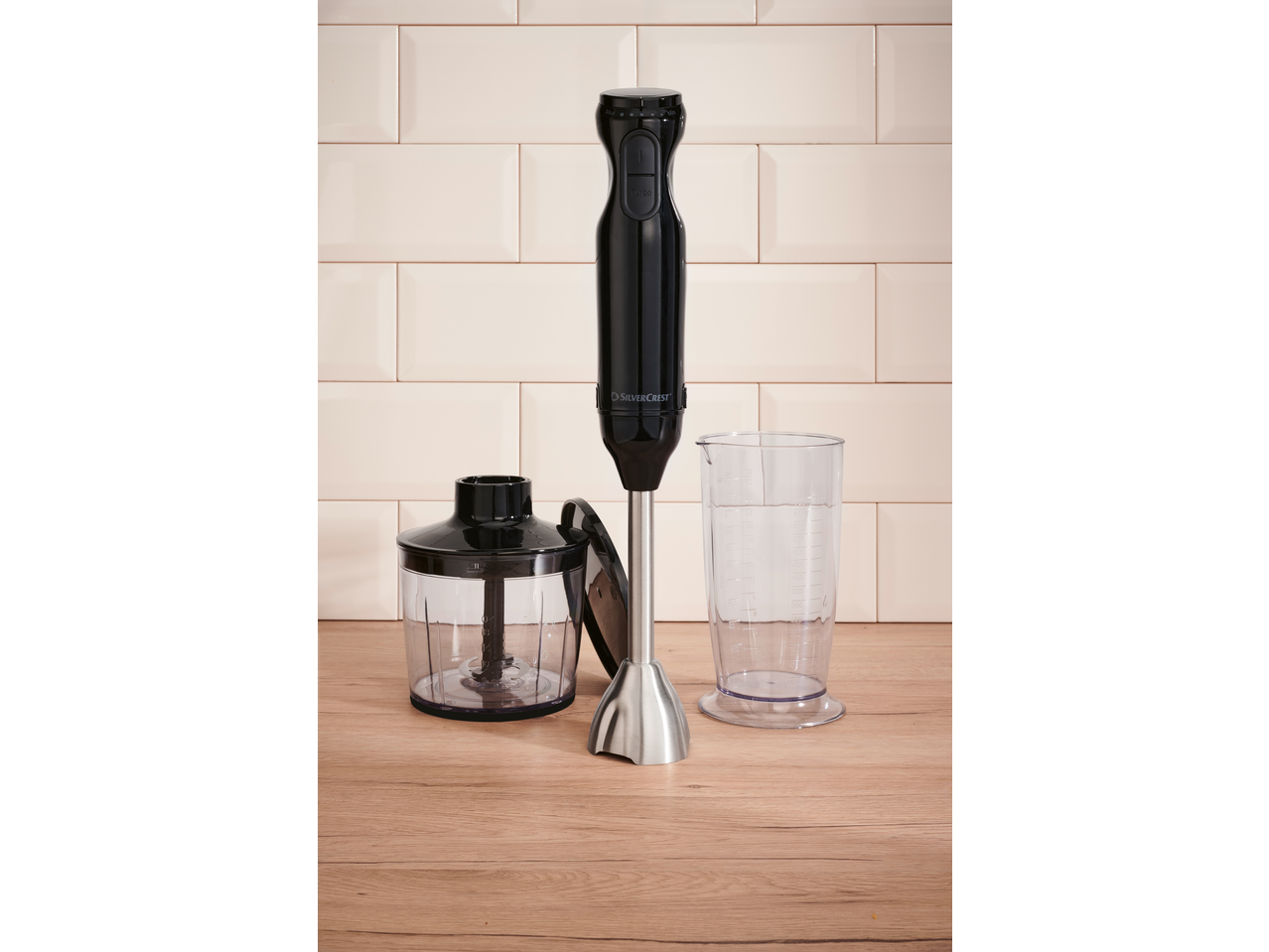 Black immersion blender with a chopper attachment and a measuring cup on a wooden surface.