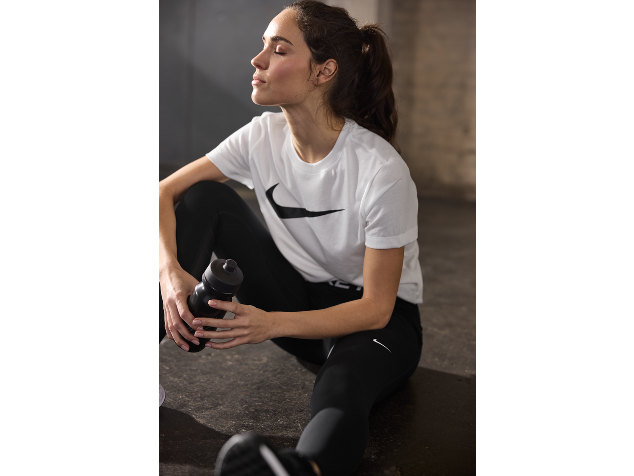 Woman in white t-shirt and black leggings holding a water bottle, resting after a workout.