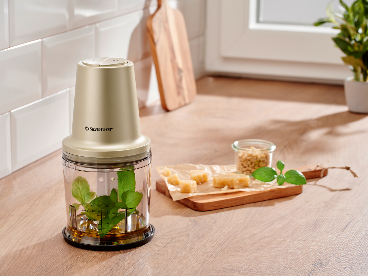 Чопър с босилек и маслиново масло, до него пармезан и кедрови ядки на кухненски плот.