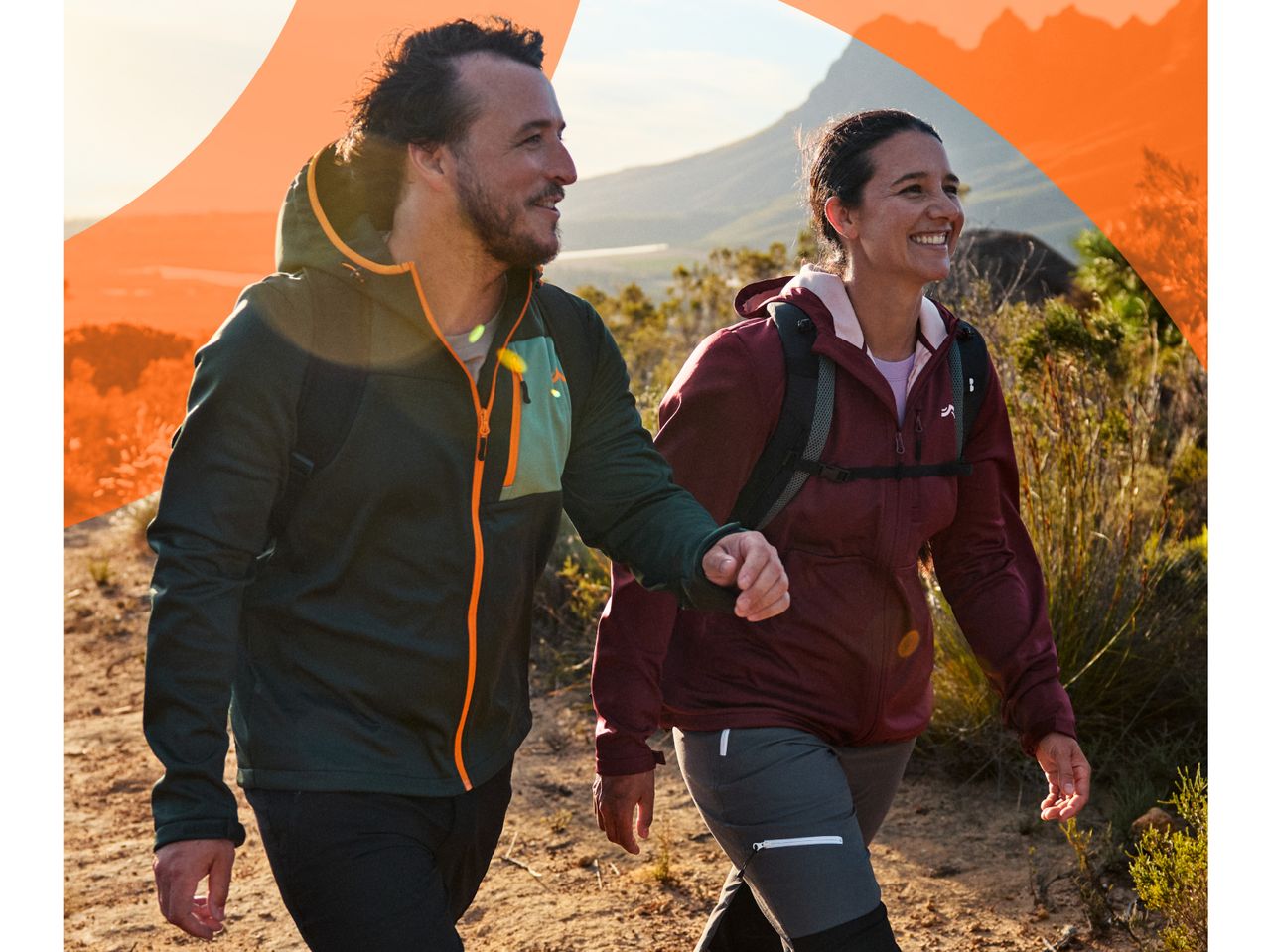 Two hikers in outdoor jackets and pants walk on a trail with mountains in the background.