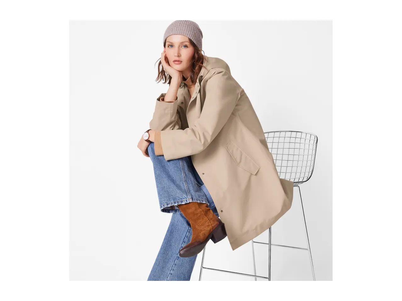 Woman in a beige raincoat, blue jeans, and brown boots, sitting on a chair.